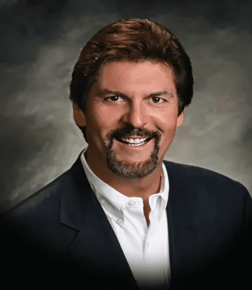 Professional headshot of a smiling man with brown hair and a beard in a suit.